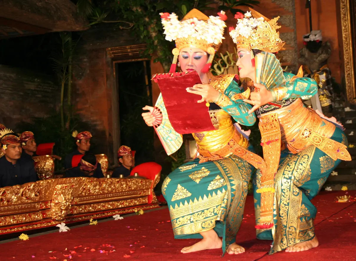 Two Legong dancers in gold and turquoise costumes and flower headdresses on a red-carpeted stage, with a gamelan ensemble seated behind them