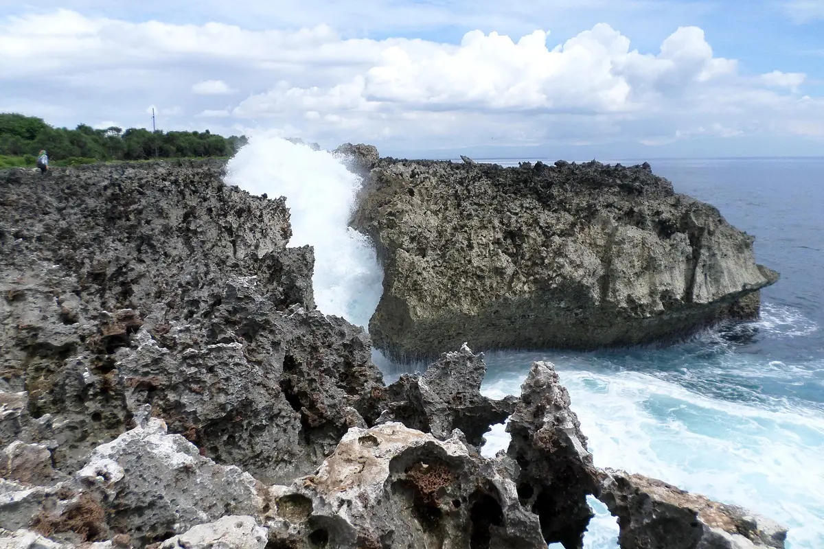 A wave breaking against jagged limestone rock at Water Blow Point in Nusa Dua, throwing spray up the cliff beside a blue sea